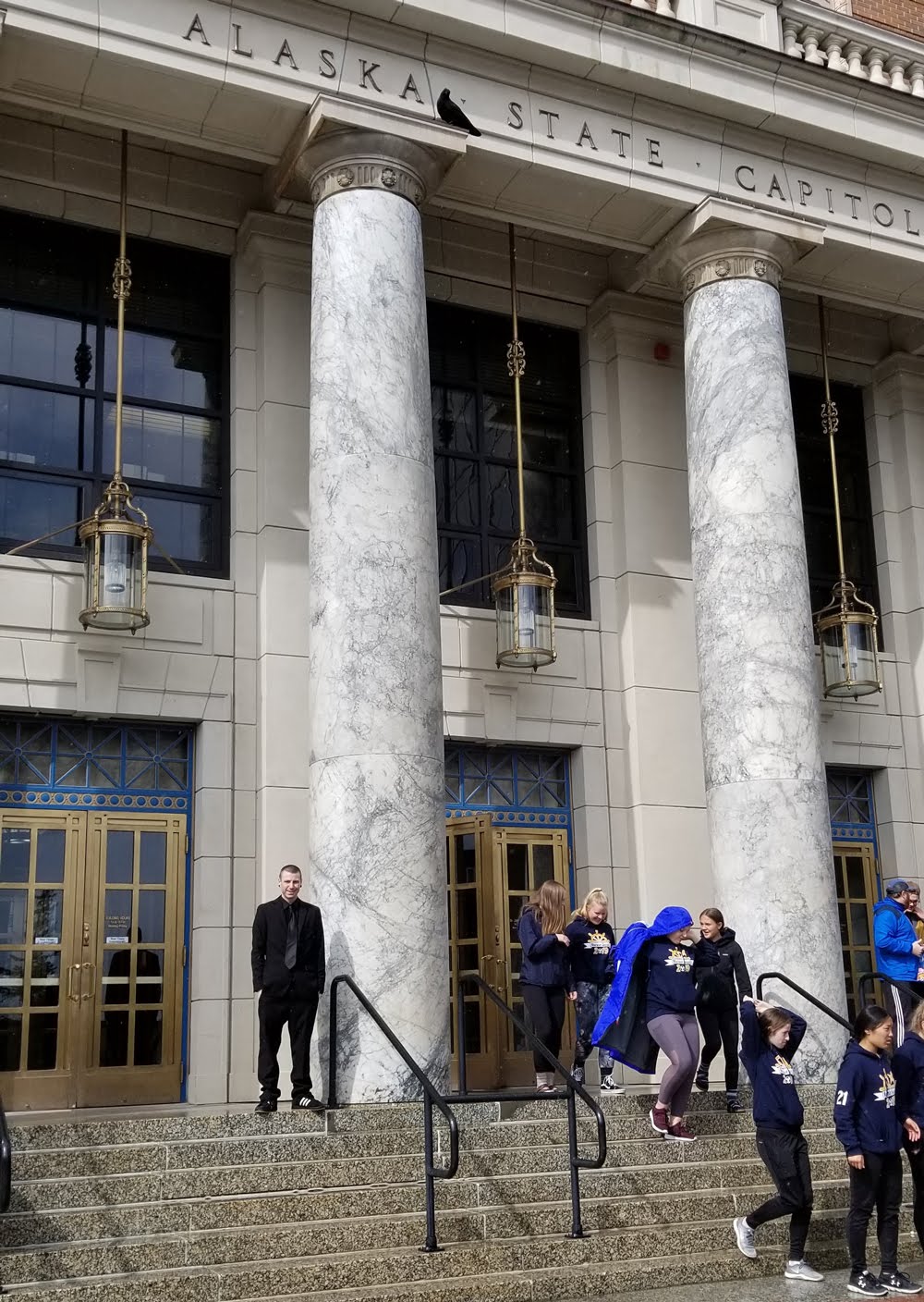 Chance Trahan in an all black suit on the steps in front of the Alaska State Capitol building with a raven perched on the top of the column right above him